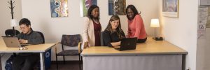 Three women gather around a laptop at one desk, while at the other a man is working on his laptop